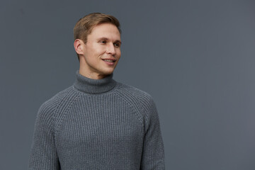 Smiling man wearing gray turtleneck sweater looking to the side against a plain gray background....