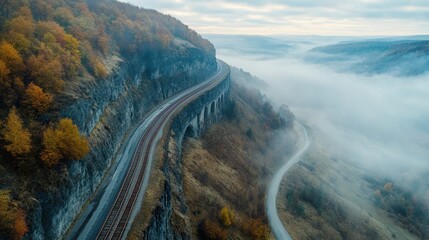 High-angle view of a winding road and railway track alongside a cliff face, shrouded in morning mist. Autumn foliage