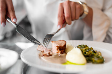 Close-up of hands cutting cooked meat on a white plate with mashed potatoes and green vegetables, depicting a gourmet dining experience.