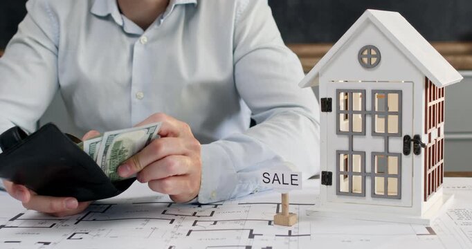 Man in white shirt opens leather wallet and pulls out bundle of dollar bills. Miniature house model stands on table with drawing. Businessman prepares for negotiations
