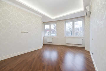 An empty room featuring smooth wooden flooring and bright natural light pouring in through large windows. The walls have a light decorative pattern, creating a modern ambiance.