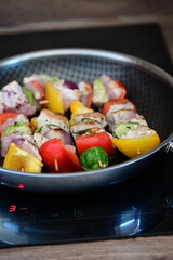 Raw chicken skewers with colorful vegetables on a black frying pan, set against the background of meal preparation