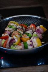 Raw chicken skewers with colorful vegetables on a black frying pan, set against the background of meal preparation