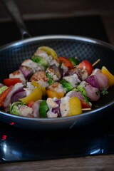Raw chicken skewers with colorful vegetables on a black frying pan, set against the background of meal preparation