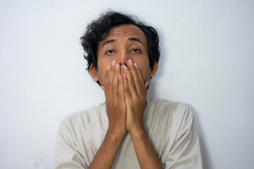 A young Indonesian man covering his mouth with both hands, expressing shock or disbelief. Perfect...