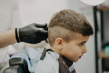 Smiling little boy sitting at barber shop, while hairdresser styling his hair.