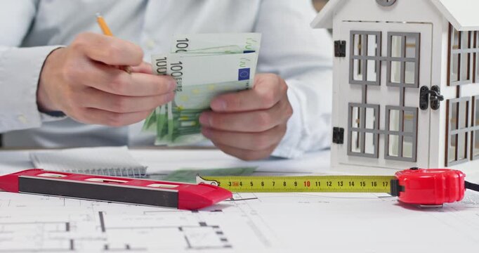 Man in white shirt counts euro banknotes and writes total amount in small notebook using pencil. Office worker sits at desk and calculates cost of building house