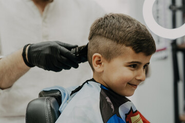 Smiling little boy sitting at barber shop, while hairdresser styling his hair.