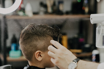 Smiling little boy sitting at barber shop, while hairdresser styling his hair.