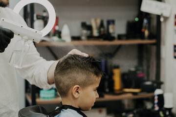 Smiling little boy sitting at barber shop, while hairdresser styling his hair.