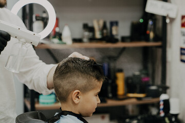 Smiling little boy sitting at barber shop, while hairdresser styling his hair.