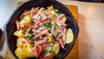 Rustic Fried Potatoes with Bacon and Herbs in Cast Iron Pan. Sliced fried potatoes topped with crispy bacon and herbs, served hot in a cast iron skillet on a wooden board.