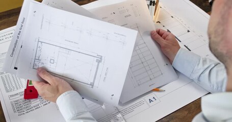 Experienced man architect sits at desk and examines sketches of different apartment rooms. Office worker in shirt pays close attention to details in bedroom layout - Powered by Adobe