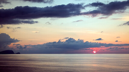 Tranquil Ocean Sunset with Dramatic Clouds. Vibrant sunset over a calm ocean with dark clouds and colorful reflections on the water near a rocky coastline.