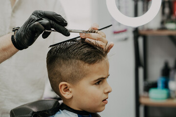 Smiling little boy sitting at barber shop, while hairdresser styling his hair.