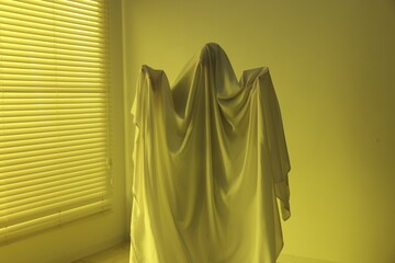 Creepy ghost. Man covered with white sheet indoors in yellow light
