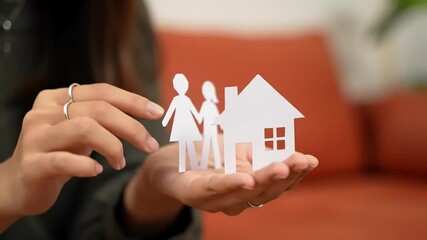 Woman's hands hold paper family and house; orange couch background; homeownership concept
