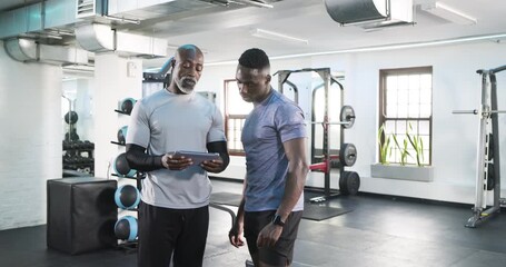 African American male trainer and client tapping tablet, planning workout and fist bumping in gym