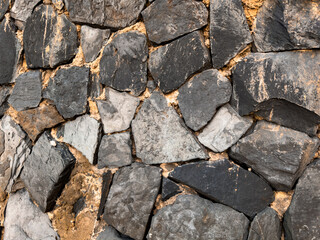 A rough stone wall made of various sized grey and black rocks, with orange-brown mortar between them, creating a textured and earthy pattern.