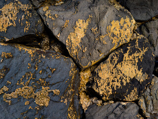 A close-up view of dark, coarse rocks with vibrant orange-yellow patches of lichen or mineral deposits, creating a rugged and colorful natural texture.