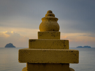 A stone monument, possibly a stupa, with a textured surface, stands against a serene backdrop of the sea and a dusky sky with a distant island.
