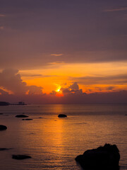 A beautiful orange and purple sunset over the calm sea, with scattered dark rocks in the foreground and a distant city skyline faintly visible on the horizon.