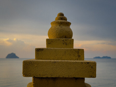 A stone monument, possibly a stupa, with a textured surface, stands against a serene backdrop of the sea and a dusky sky with a distant island.