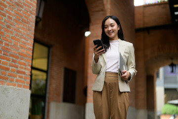 Young Asian businesswoman using smartphone and holding laptop in brick archway