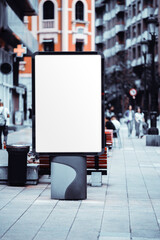 Blank vertical city light mockup with empty white advertising space on a pedestrian street, surrounded by urban architecture, benches, and blurred walking people in soft daylight