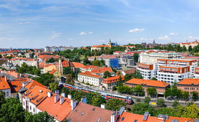 Obraz premium Panoramic view of Prague cityscape with red-tiled rooftops, residential buildings, and a commuter train passing through the urban area on a sunny summer day.
