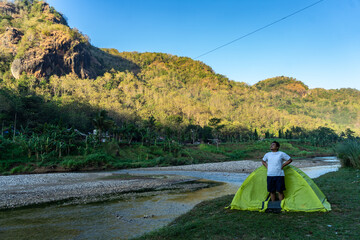 Asian man waking up by a riverside campsite, soaking in the morning view of misty hills. A peaceful...