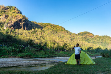 Asian man waking up by a riverside campsite, soaking in the morning view of misty hills. A peaceful...
