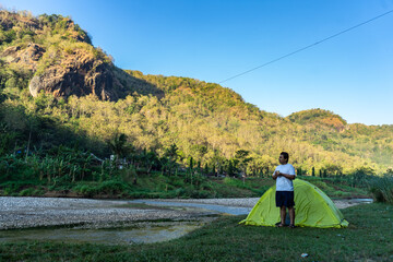 Asian man waking up by a riverside campsite, soaking in the morning view of misty hills. A peaceful...
