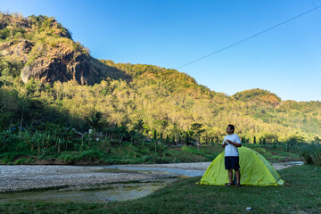 Asian man waking up by a riverside campsite, soaking in the morning view of misty hills. A peaceful...
