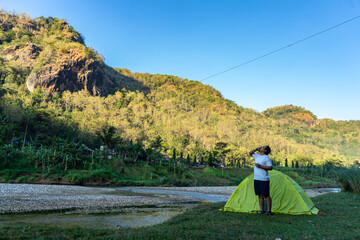 Asian man waking up by a riverside campsite, soaking in the morning view of misty hills. A peaceful...