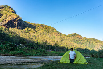 Asian man waking up by a riverside campsite, soaking in the morning view of misty hills. A peaceful...