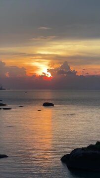 A beautiful sunset over the sea with a radiant orange and yellow sky, silhouetted rocks in the foreground, and the sun dipping below the horizon.