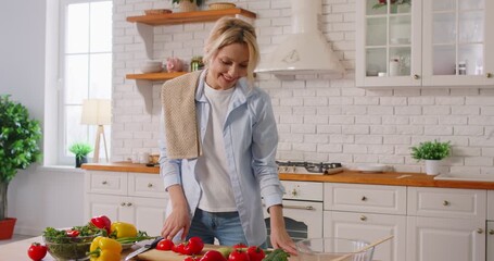 Happy middle aged European woman enjoying cooking process, prepares organic salad, chopping fresh vegetables, posing against well equipped cozy kitchen interior, keeping to healthy nutrition - Powered by Adobe