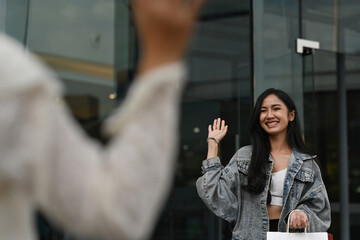 An Asian woman in a denim jacket waves goodbye to a friend with a cheerful smile after a fun shopping day outside a modern mall building