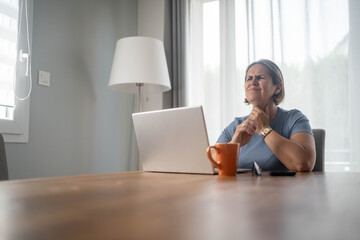 Sick mature woman with sore throat working from home on laptop, suffering from flu or allergy in...