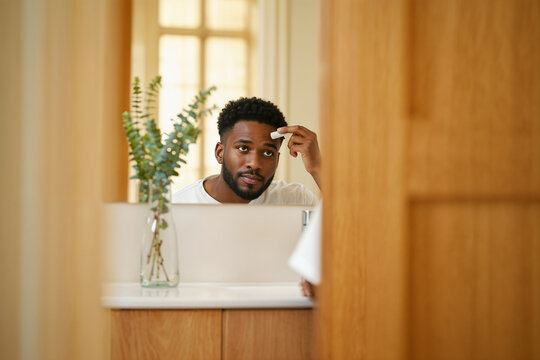 Man's skincare routine. A young African American man looking in the mirror while applying a facial product to his forehead in a modern bathroom.