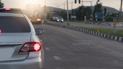Rear side view of silver car wtih turn on brake light. Stop the long queue from the traffic light at the intersection. Center of raod with island of green grass. Car across the road in the morning.