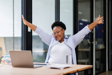 Excited cheerful Black african american woman using laptop computer on sofa at workplace, getting good news, feeling joy, dancing with hands, singing, laughing, making winner gesture, happy to win pri