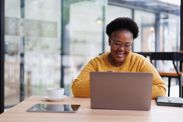 Cheerful freelancer working on a laptop in a modern coworking space, savoring a cup of coffee while jotting down notes in her notebook and browsing on her tablet