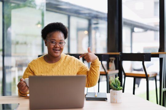 Young african american woman is pointing at herself with her finger while holding a cup of coffee and working on a laptop in a cafe, showing self-confidence and pride in her work