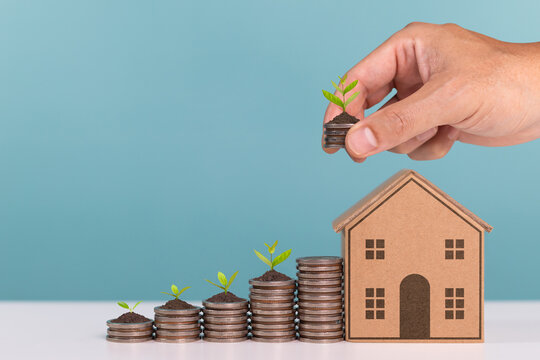 A hand places a small stack of coins with a growing plant next to a house model, symbolizing sustainable financial investment, green finance, and home savings for future growth