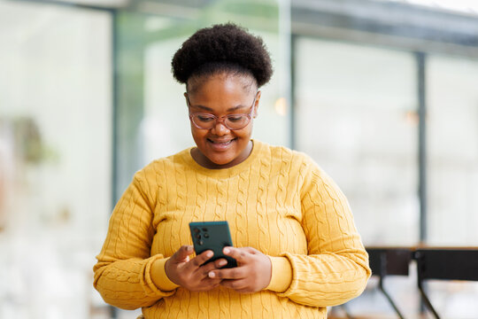 Happy attractive African American business lady using Internet technology for job in co-working space, sitting at laptop, holding mobile phone.
