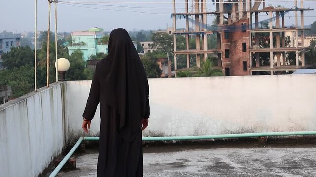 A confident Bengali woman in a black burqa and hijab stands on the edge of a rooftop, looking out at the cityscape. The video is shot from the back, highlighting her peaceful posture.