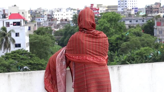 Bengali Muslim woman in salwar kameez with head veil, standing on a rooftop in the morning sunlight, enjoying fresh air and city view. Back view.