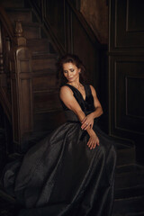 A woman in a stunning black gown poses gracefully on an old wooden staircase. Her expression is serene and confident, capturing the beauty of the elegant surroundings.